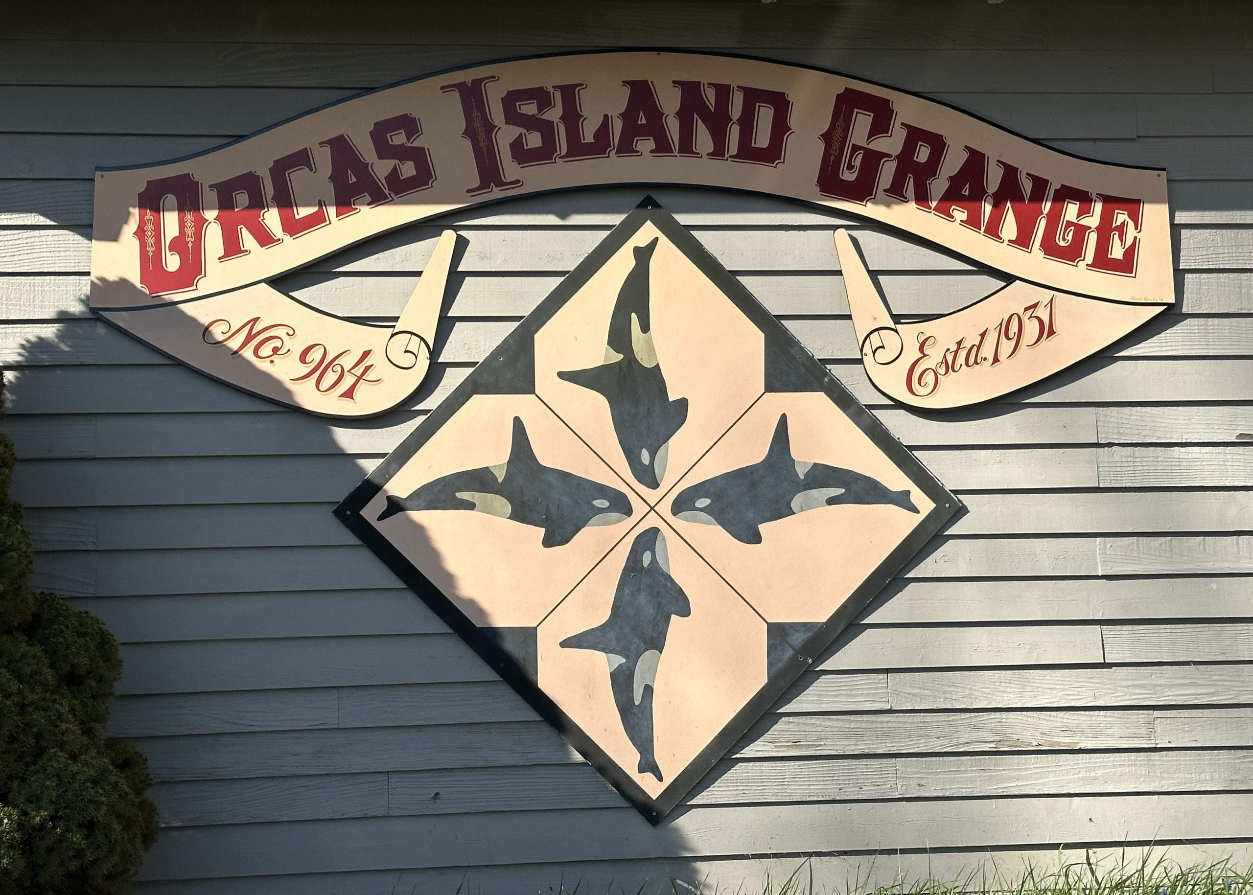 Orcas Island Grange sign mounted on a gray wooden wall, featuring four orca whales in a diamond pattern and decorative lettering reading ‘Orcas Island Grange No. 964 Est. 1931.